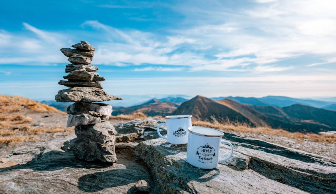 Two enamel mugs with inspirational quotes sit on a rocky mountaintop beside a stack of balanced stones, with scenic mountain peaks in the background under a bright blue sky.