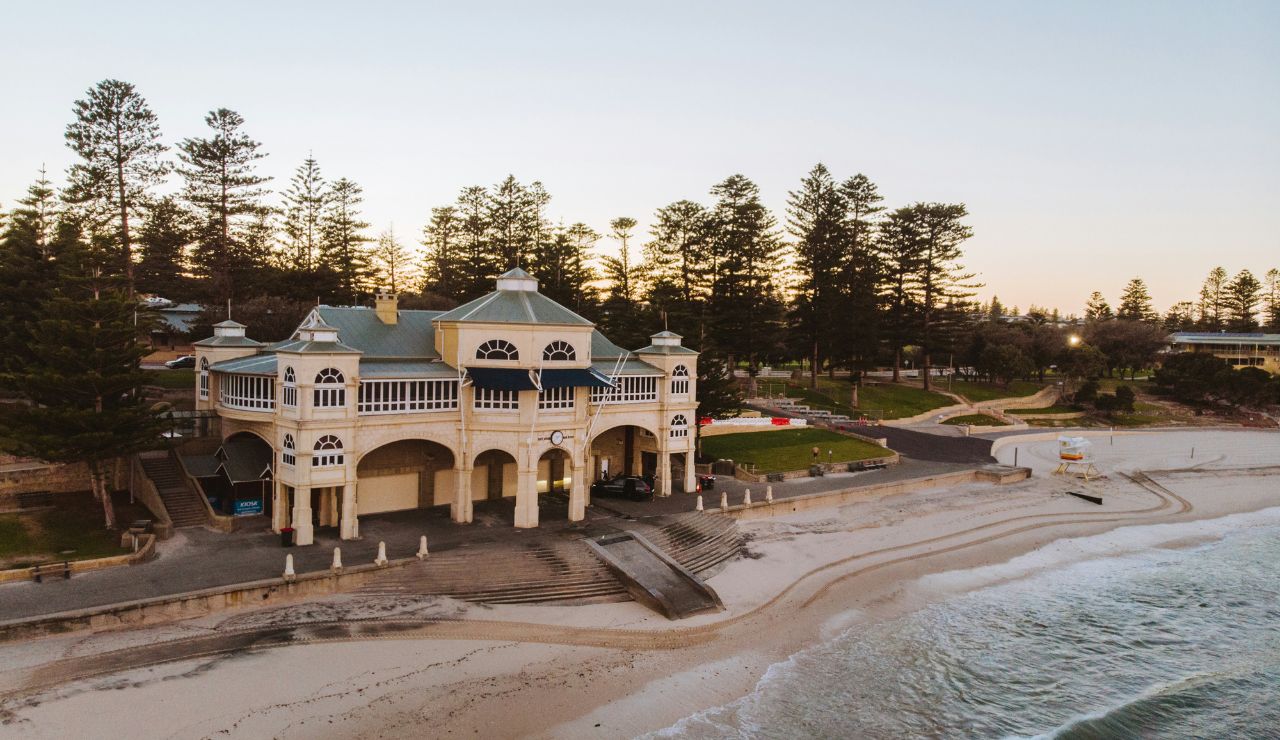 Elegant beachfront pavilion with arched architecture overlooking a sandy shore, surrounded by pine trees and gentle ocean waves at sunset.