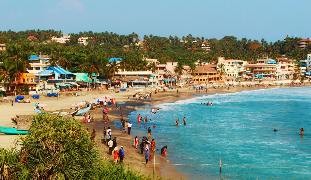 People swim and walk along a busy beach bordered by colorful buildings and palm trees under a clear sky.