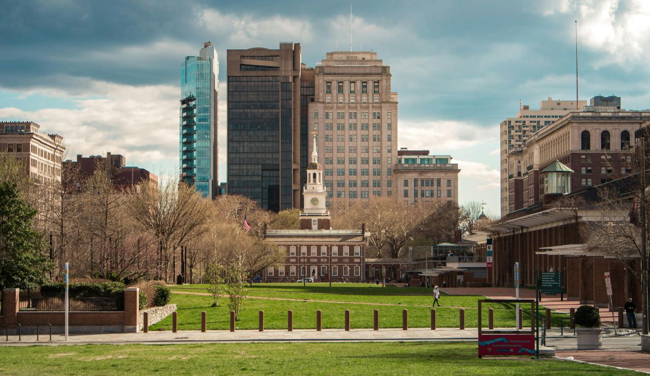 Independence Hall stands before modern office buildings, framed by trees and a large green lawn under a partly cloudy sky.