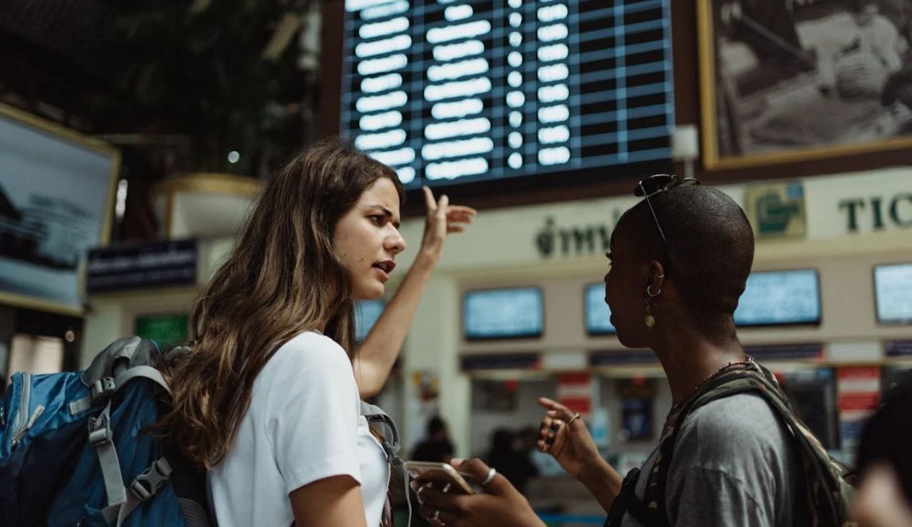Two travelers with backpacks discussing in front of a departure board at an Airport.