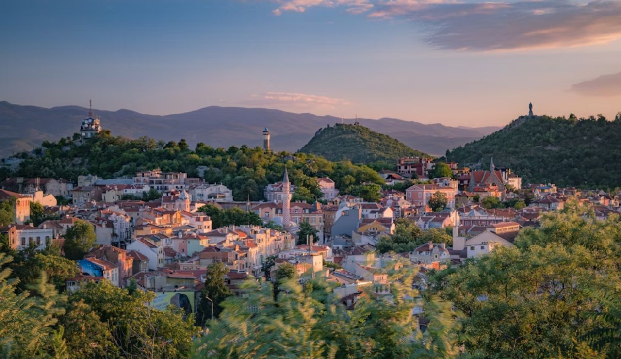 Scenic view of a European town nestled among green hills during sunset, with historic buildings and distant mountain ranges.