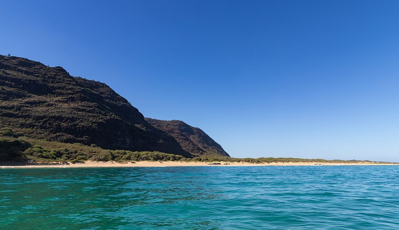 Coastal view of a remote beach with turquoise water and dark, rocky cliffs under a clear blue sky.