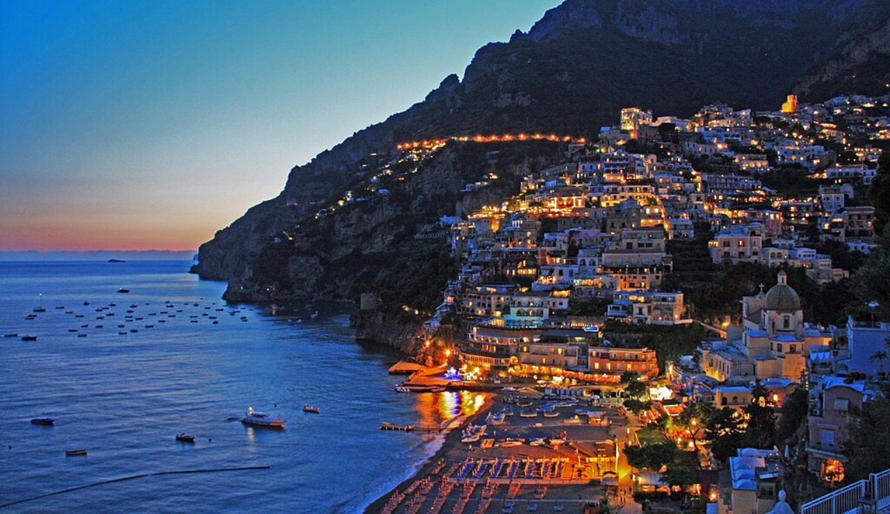 Colorful buildings of Positano glowing at dusk along the Amalfi Coast, with boats floating on the calm sea and beach umbrellas lined up below.