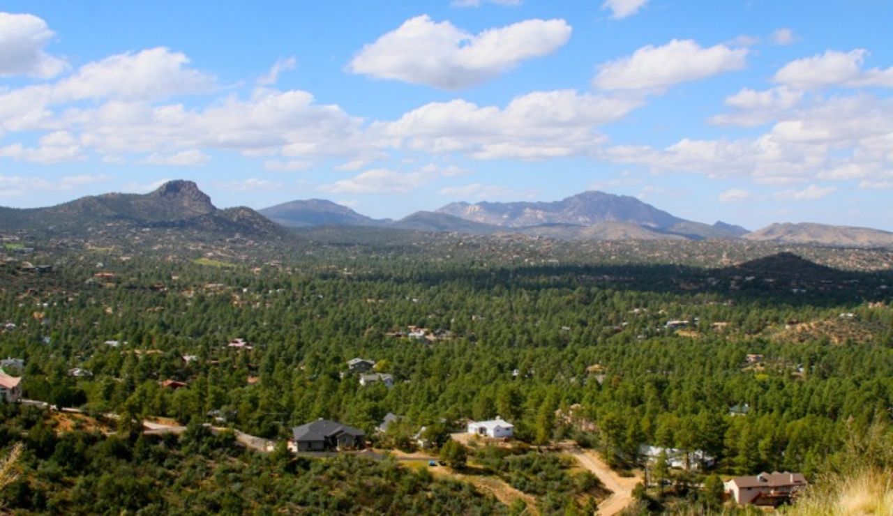 A scenic view of Prescott, Arizona, shows scattered homes nestled among pine forests with mountains rising in the distance under a bright, cloud-dotted sky.