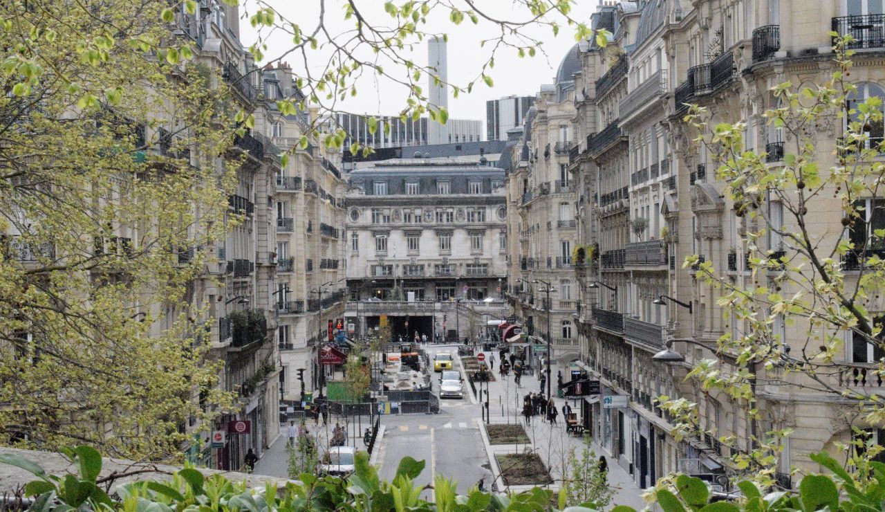 A bustling city street features classic European buildings, busy traffic, pedestrians, and leafy trees framing the scene.