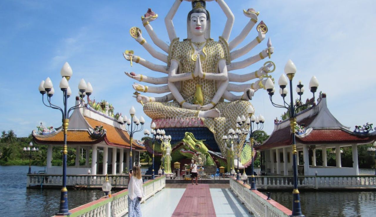 ourist walking toward a large, multi-armed Guanyin statue at Wat Plai Laem temple in Thailand, surrounded by water and traditional architecture.