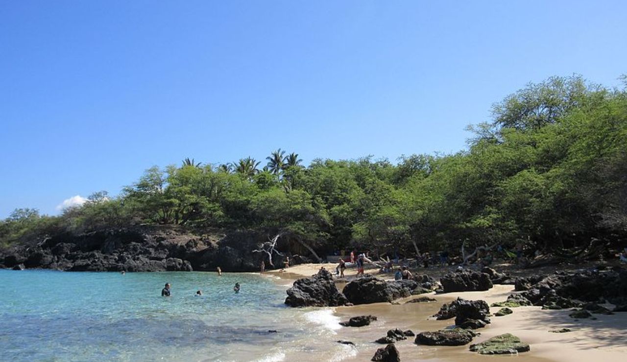 Small rocky beach with clear turquoise water, surrounded by lush trees and visitors enjoying the shoreline.