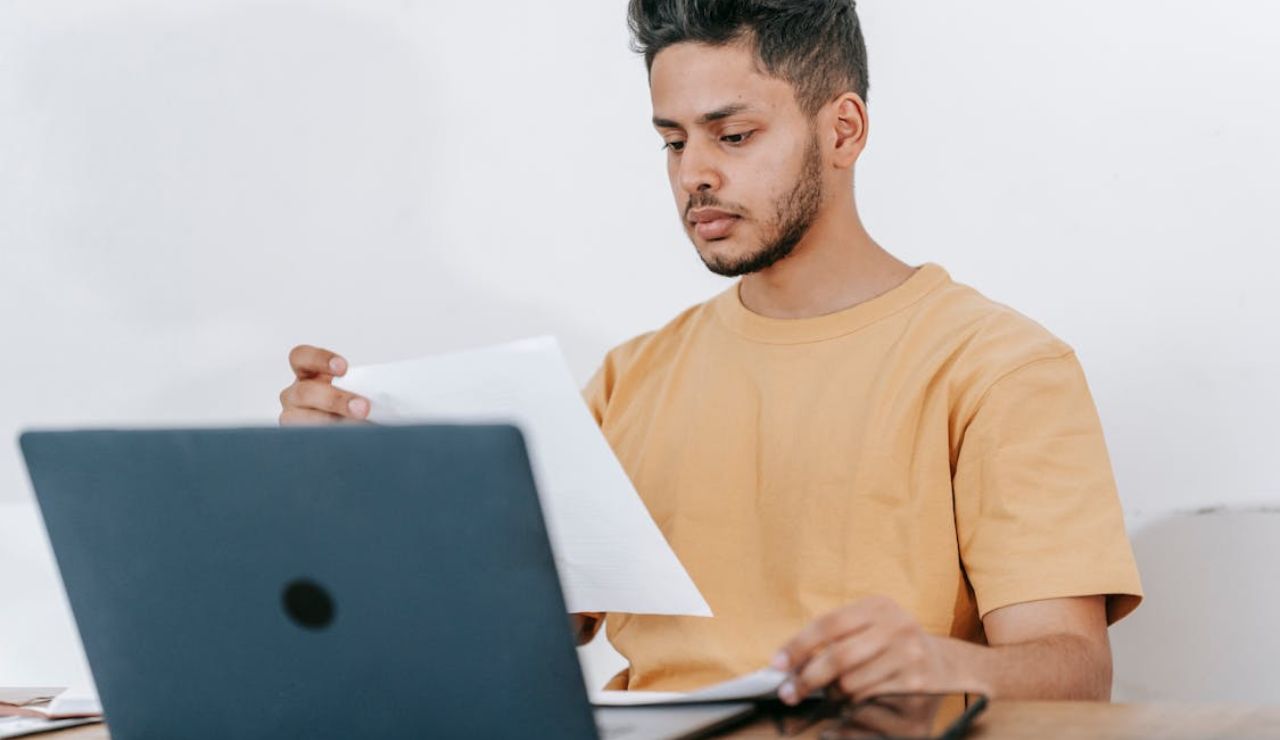 Man in yellow shirt reading papers in front of a laptop while researching.