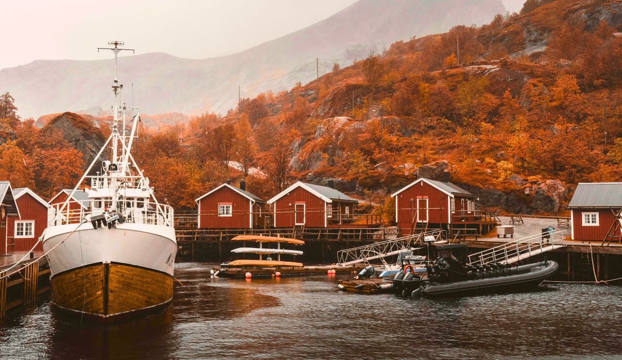 A white boat is docked by small red stilted cabins beside calm water, with autumn trees and misty mountains in the background.