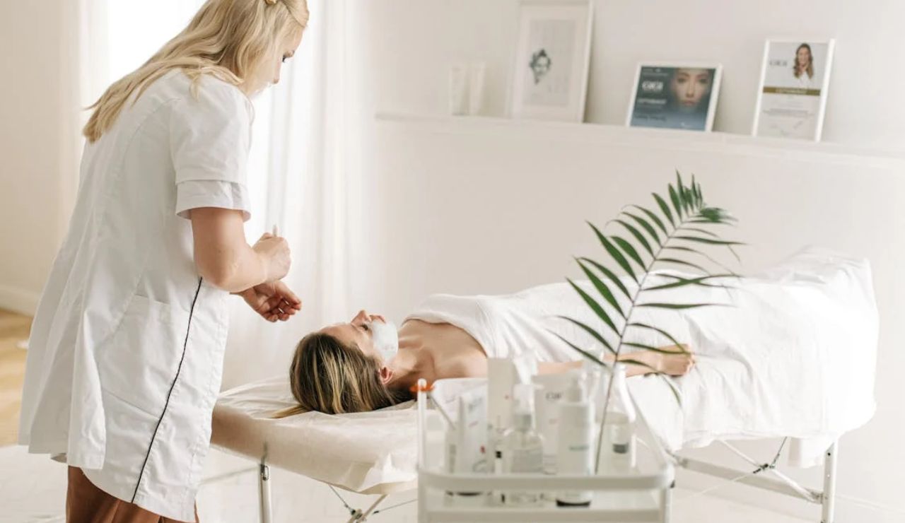 A woman receiving a facial treatment in a spa setting, with a therapist applying a mask while the room is filled with skincare products.