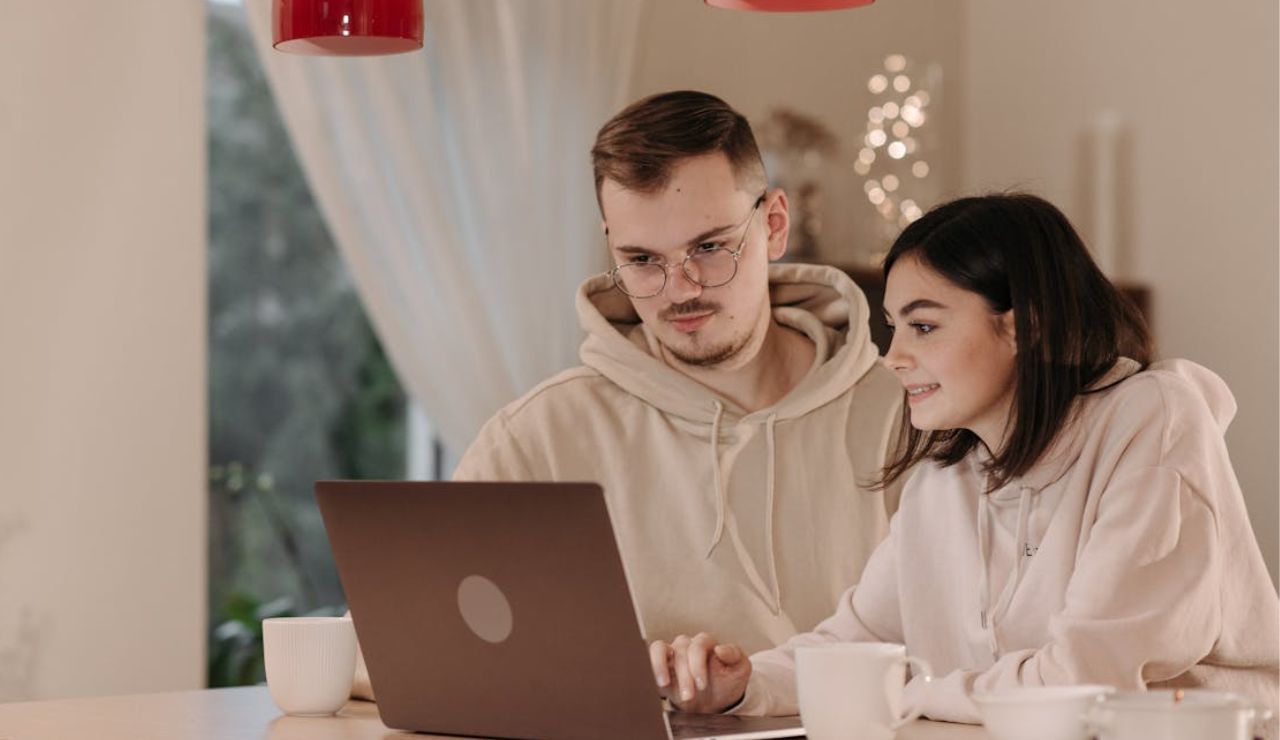 Couple sitting together at a table, looking at a laptop screen and smiling in a cozy indoor setting.