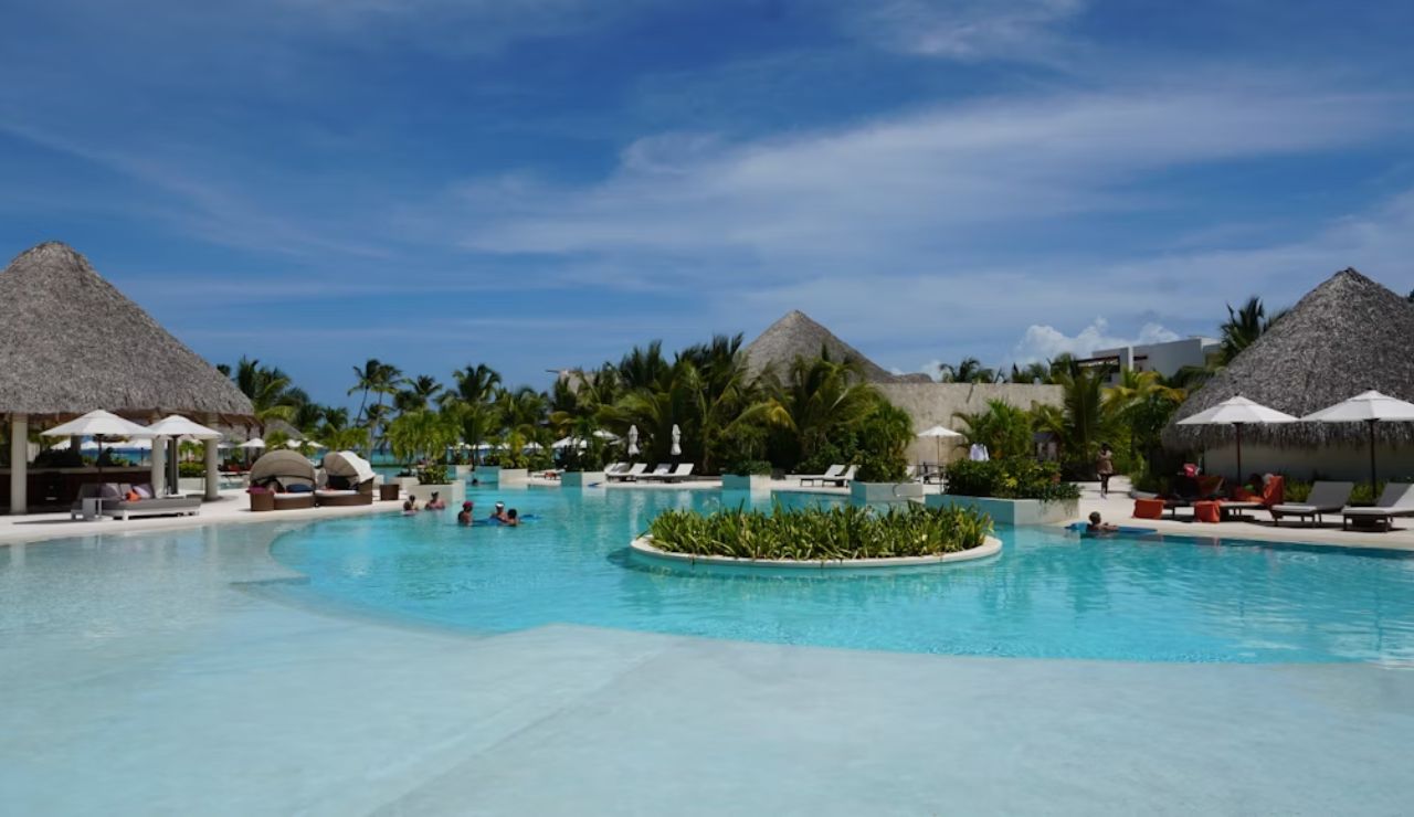 Tropical resort pool surrounded by palm trees, lounge chairs, and thatched-roof cabanas under a sunny blue sky.