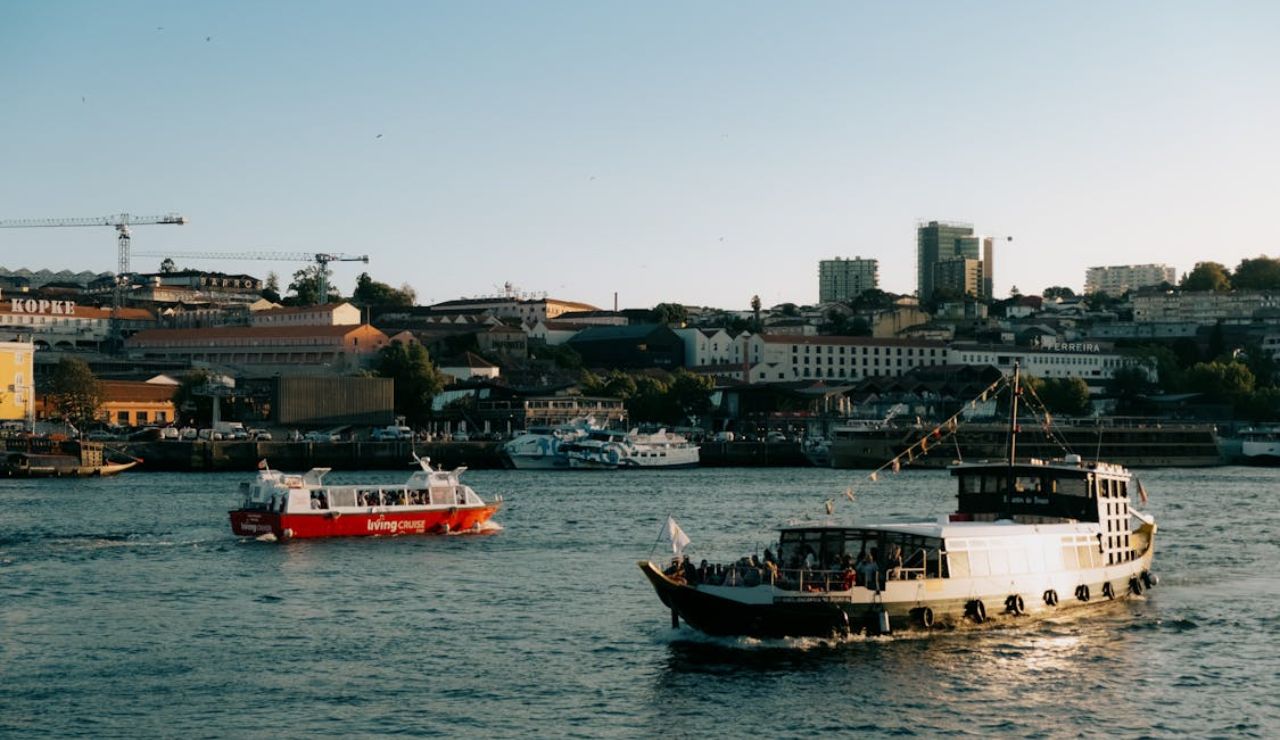 Two river cruise boats sailing near a city waterfront with buildings and cranes in the background.