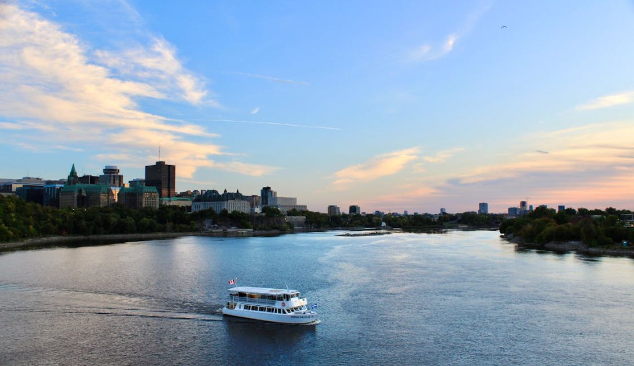 White river cruise boat sailing on a calm river at sunset with a city skyline in the background and a Canadian flag on the boat.