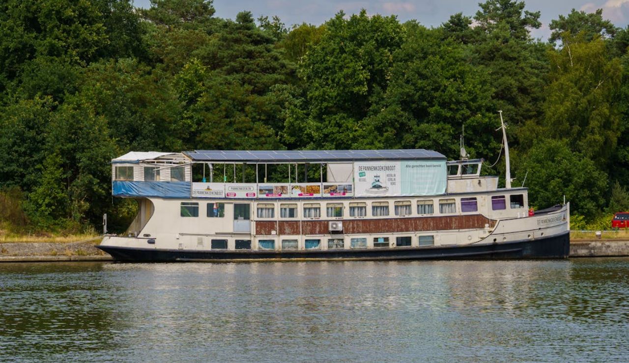 A docked riverboat restaurant named "De Pannenkoekenboot" with dining space on the upper deck, surrounded by lush green trees.
