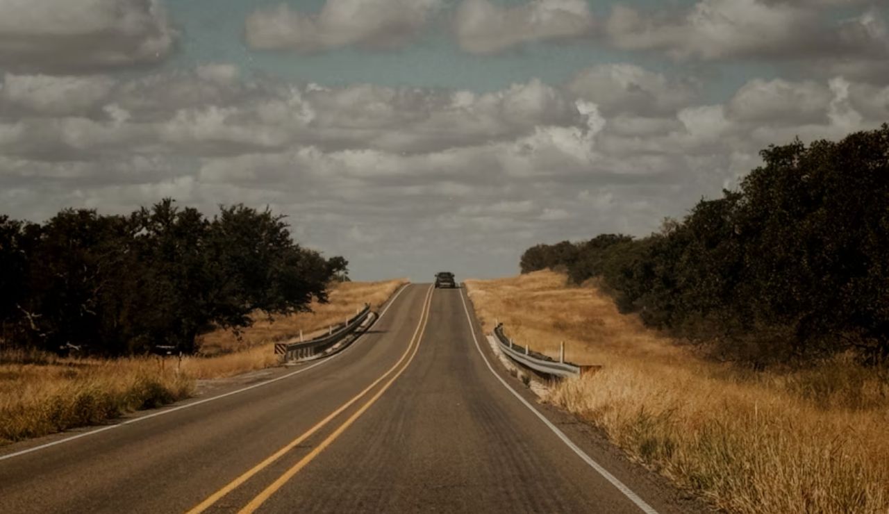 Car driving on an empty rural highway surrounded by fields and trees.