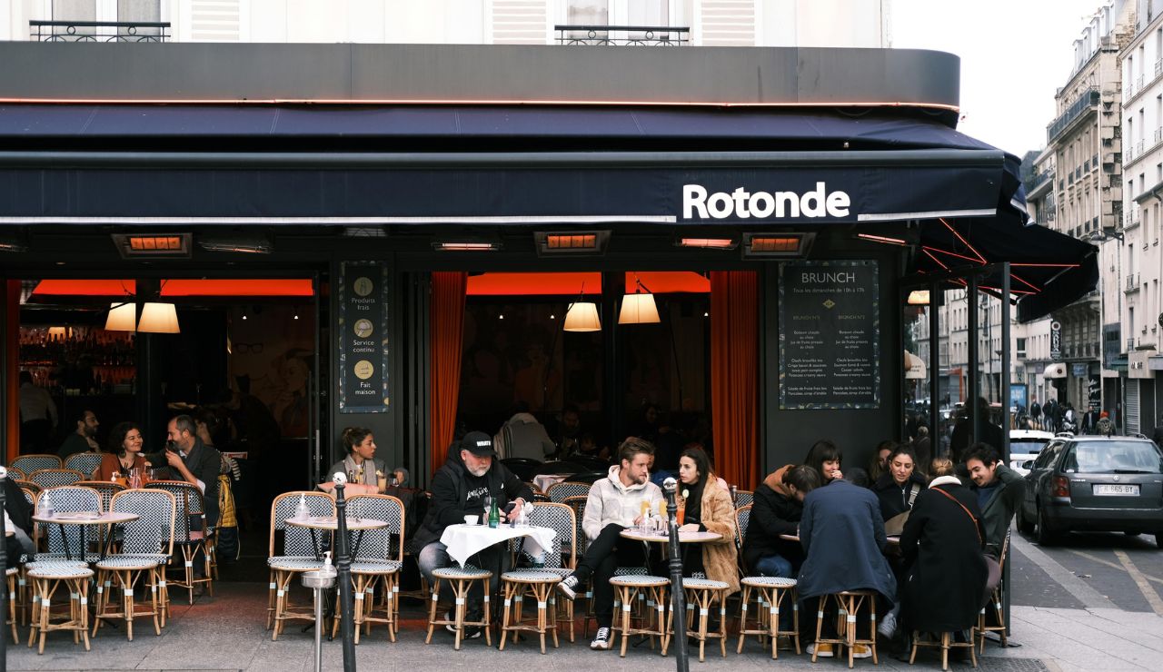 People socialize at outdoor tables outside Rotonde café on a city street.