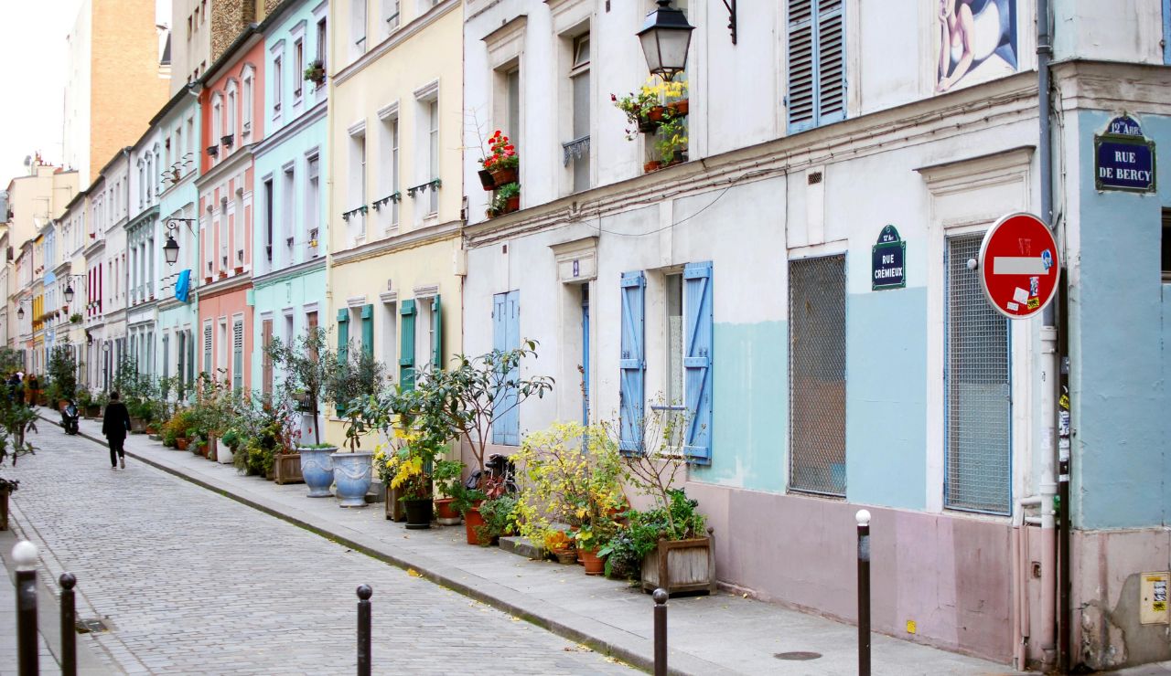 A narrow cobblestone street lined with colorful buildings, potted plants, shuttered windows, street signs, and a lone figure walking away.