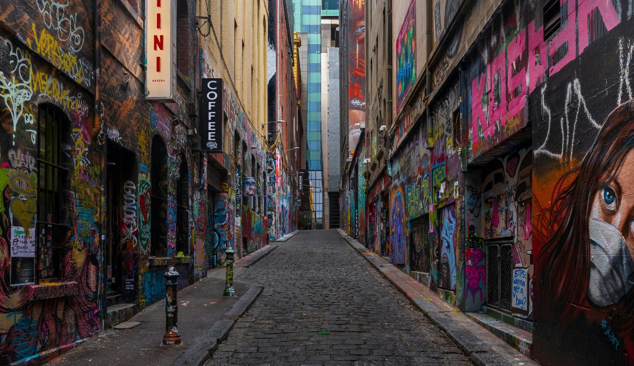 A narrow cobblestone alley features colorful graffiti, a coffee shop sign on the left, and a large masked face mural on the right.