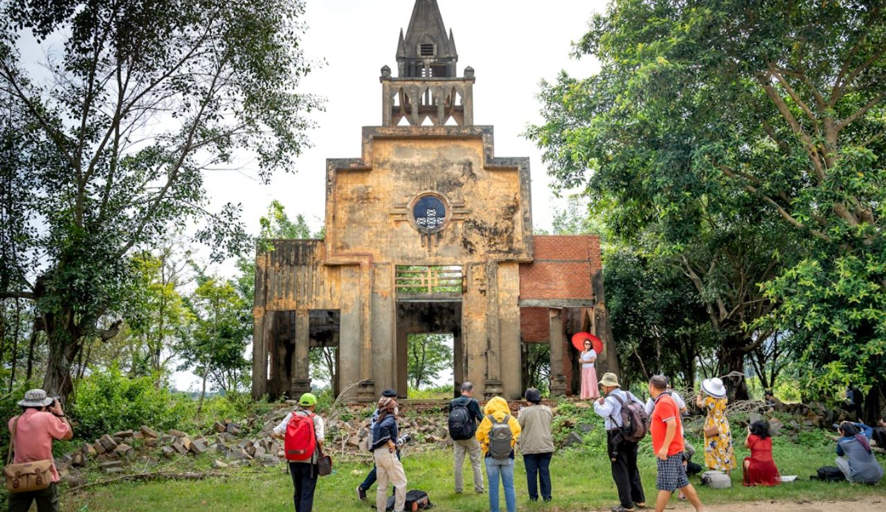 Group of tourists and photographers gathered in front of an old, weathered church ruin surrounded by lush green trees.