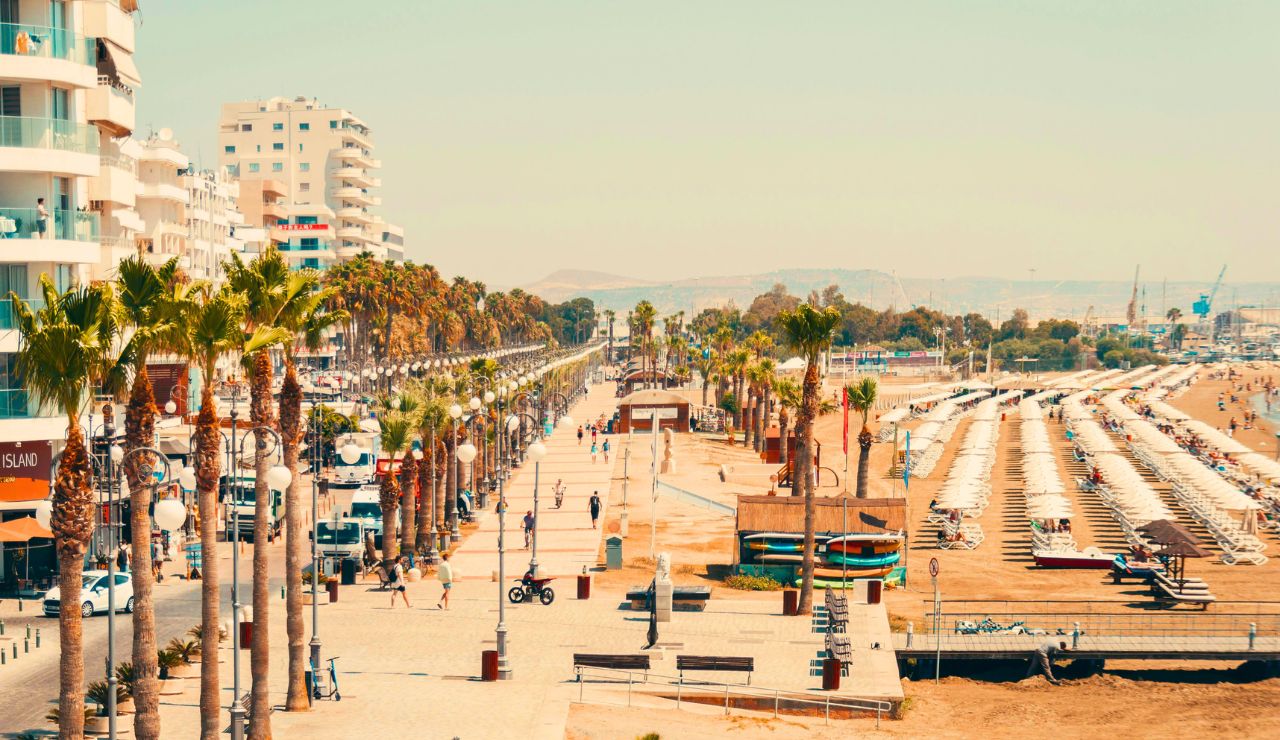 A coastal promenade bordered by palm trees features modern buildings on the left and a sandy beach with umbrellas and sunbeds on the right.