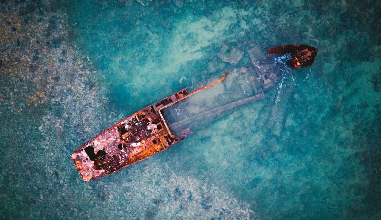 Aerial view of a rusted shipwreck submerged in clear turquoise water with a small boat nearby.