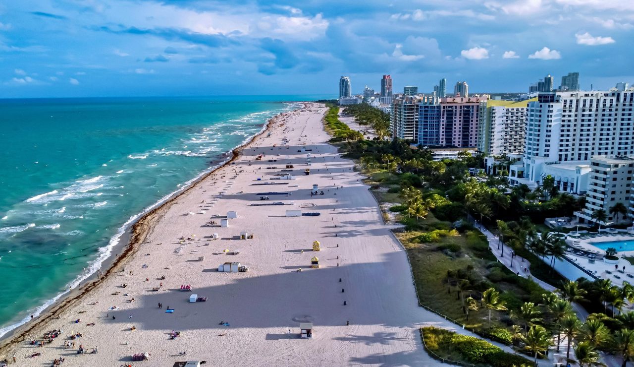 Aerial view of a sandy beach with scattered people, turquoise water, and high-rises along the shoreline beneath a partly cloudy sky.