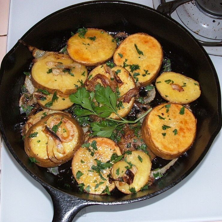 Sliced golden-brown potatoes sautéed with onions and garnished with parsley in a cast iron skillet on a stovetop.