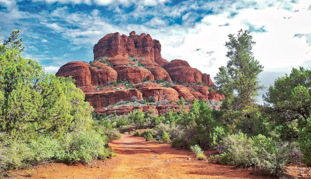 Dirt trail leading through green shrubs to the striking red rock formation of Bell Rock under a partly cloudy sky in Sedona, Arizona.