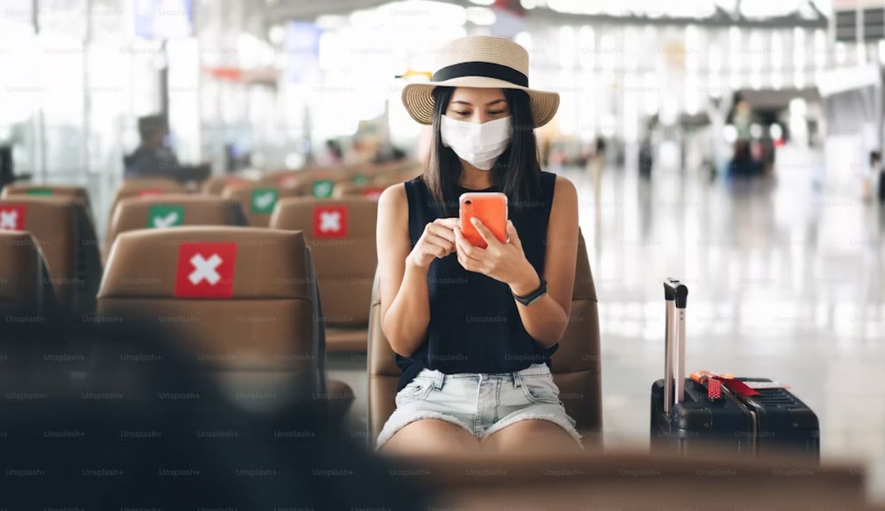 Woman wearing a face mask and hat sitting in an airport terminal using her phone, with luggage beside her and social distancing signs on seats.