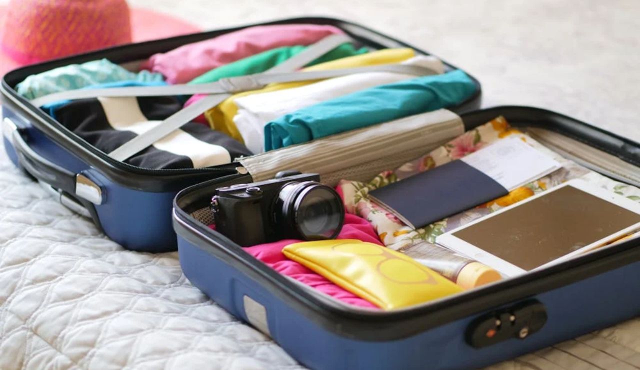 Open suitcase on a bed packed with colorful clothes, camera, passport, tablet, and travel essentials.