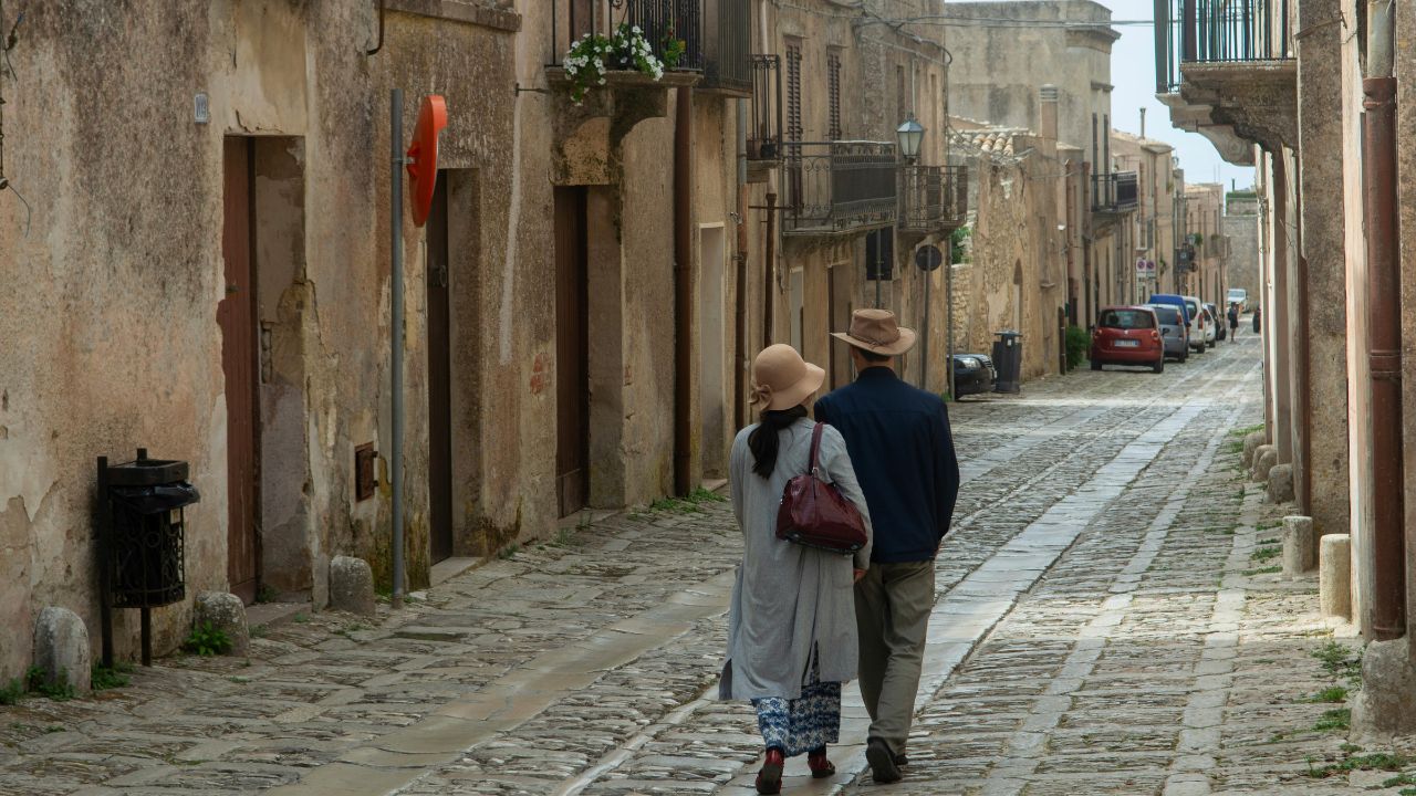 Elderly couple walking arm-in-arm down a narrow, cobblestone street lined with old stone buildings.