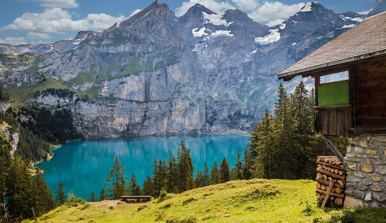 A wooden mountain cabin overlooking a turquoise lake surrounded by pine trees and dramatic snow-capped cliffs.