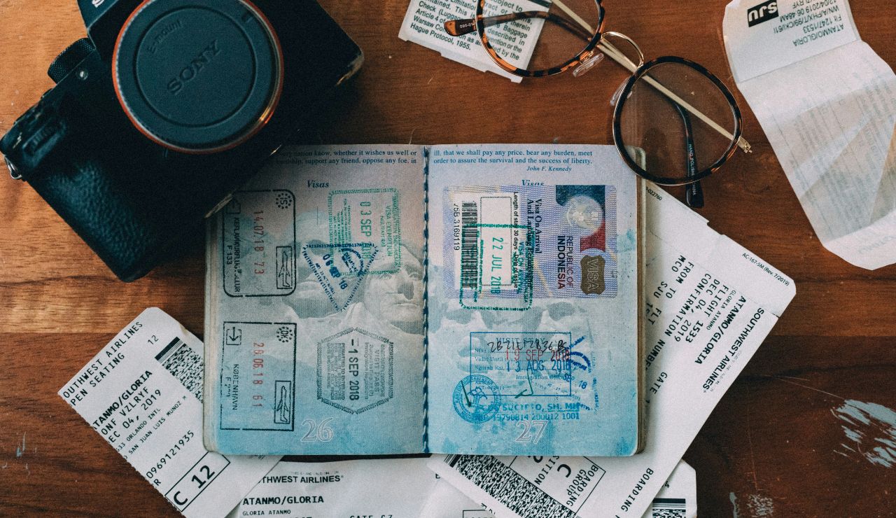Open passport with visa stamps, flight tickets, camera, and glasses on a wooden table.