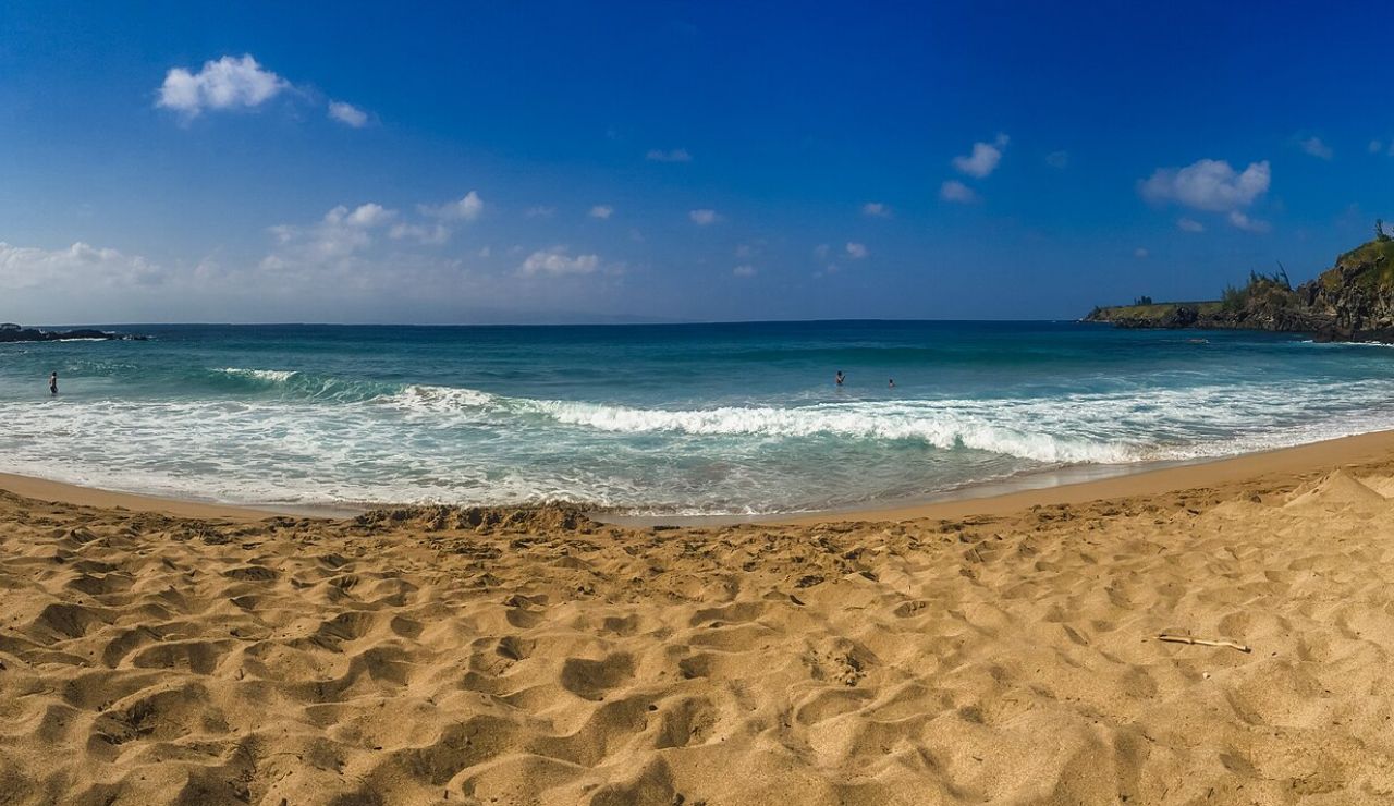 Wide sandy beach with gentle waves rolling in under a bright blue sky and scattered clouds.