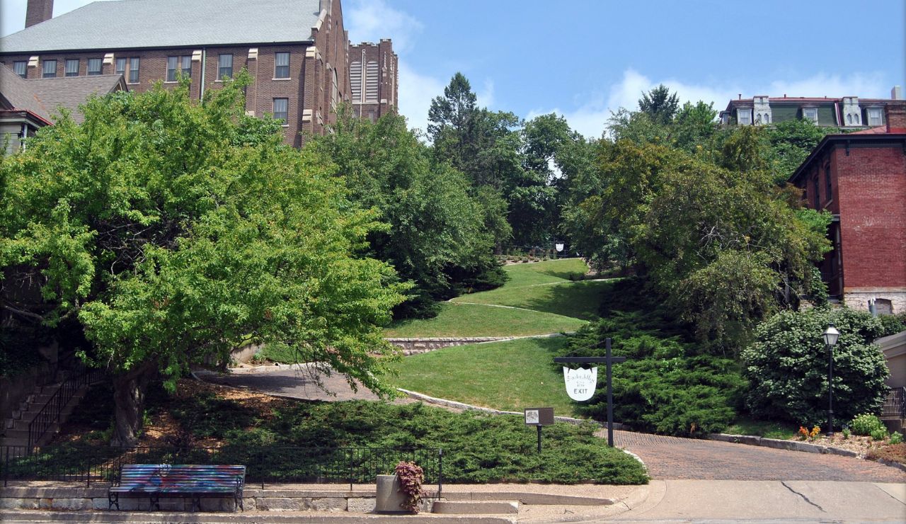 A sloped grassy path with stairs ascends between brick buildings and trees under a blue sky, with a bench and sign in the foreground.