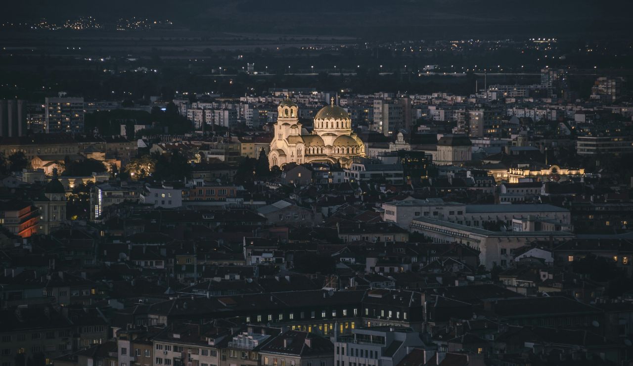 Nighttime cityscape with a brightly lit cathedral featuring golden domes, surrounded by darker urban buildings and distant lights on the horizon.