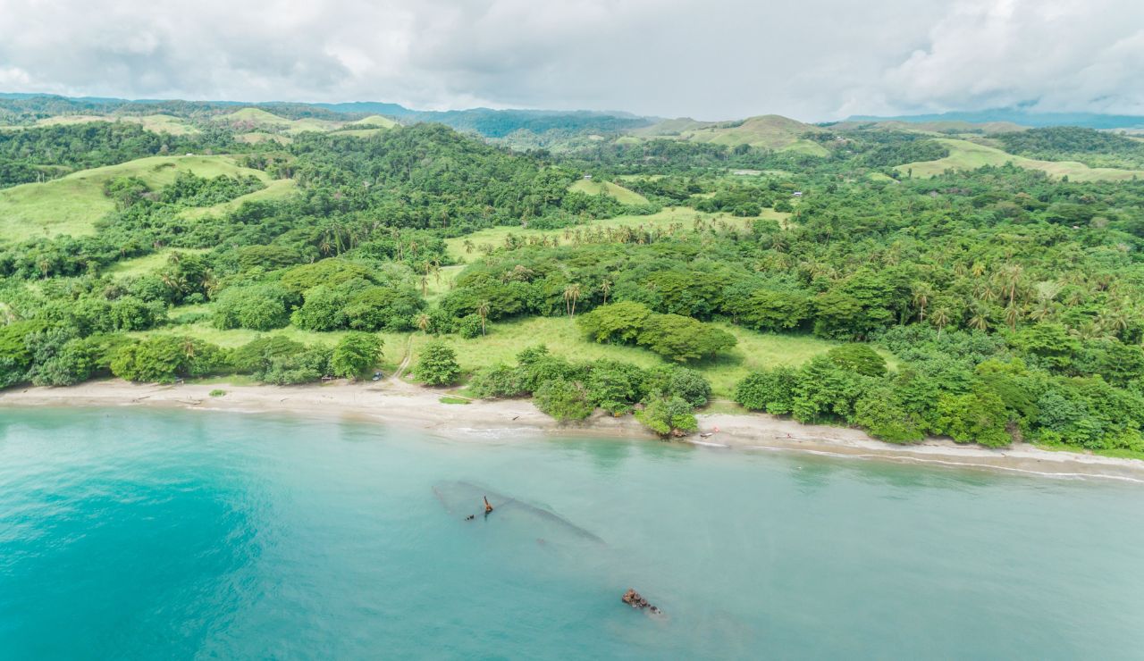 Aerial view of a lush green coastline with dense forest, sandy beach, and a partially submerged shipwreck in the clear blue water.