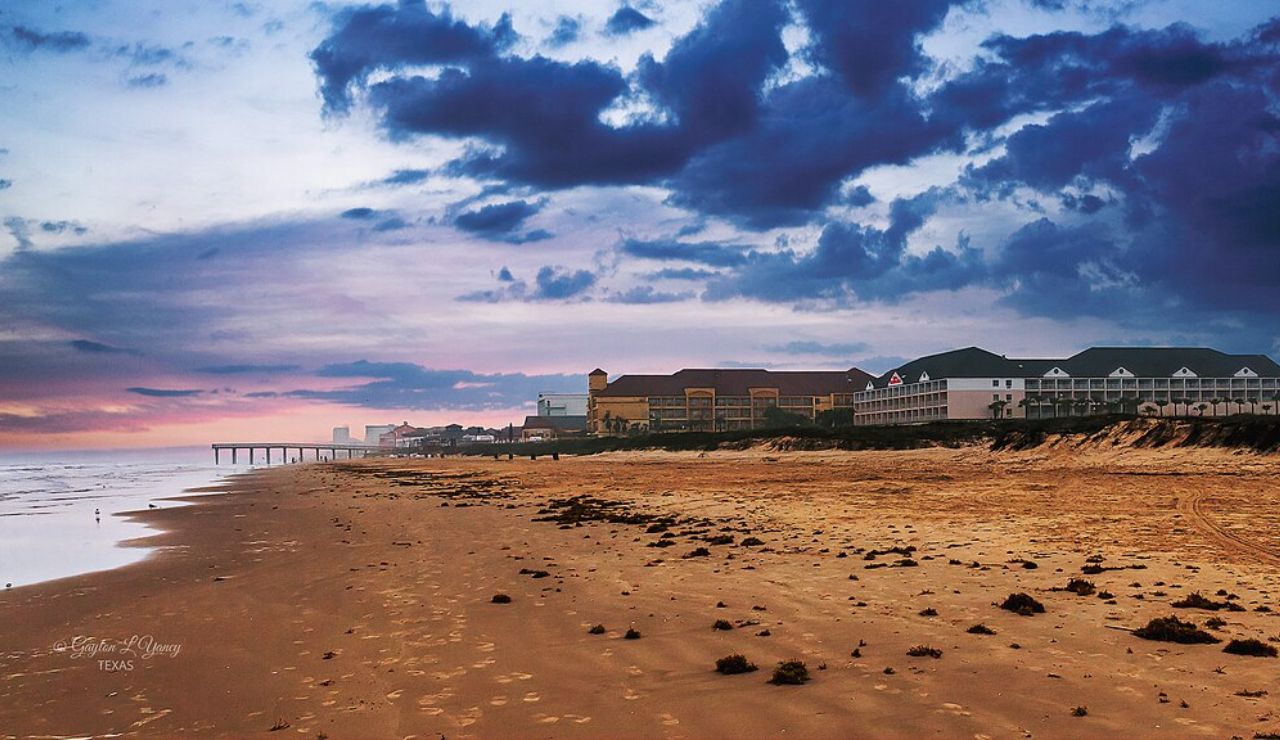 Sandy beach at sunset with buildings and a pier in the distance under a dramatic, cloud-filled sky.