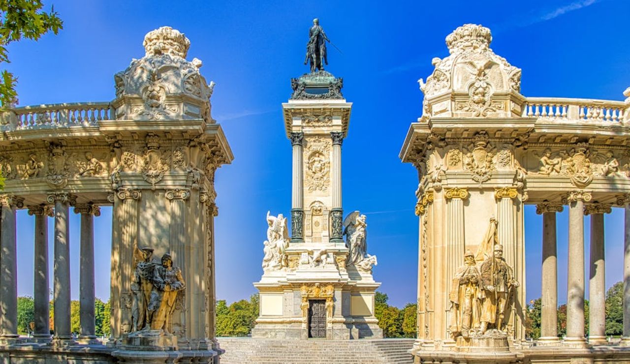A grand monument with ornate columns and statues in El Retiro Park, Madrid, featuring the statue of King Alfonso XII on a central pedestal.