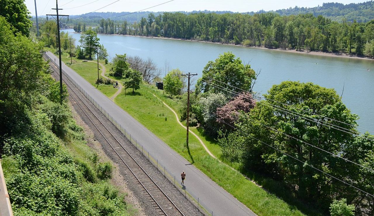 Paved riverside trail runs beside railroad tracks with trees and a river view.