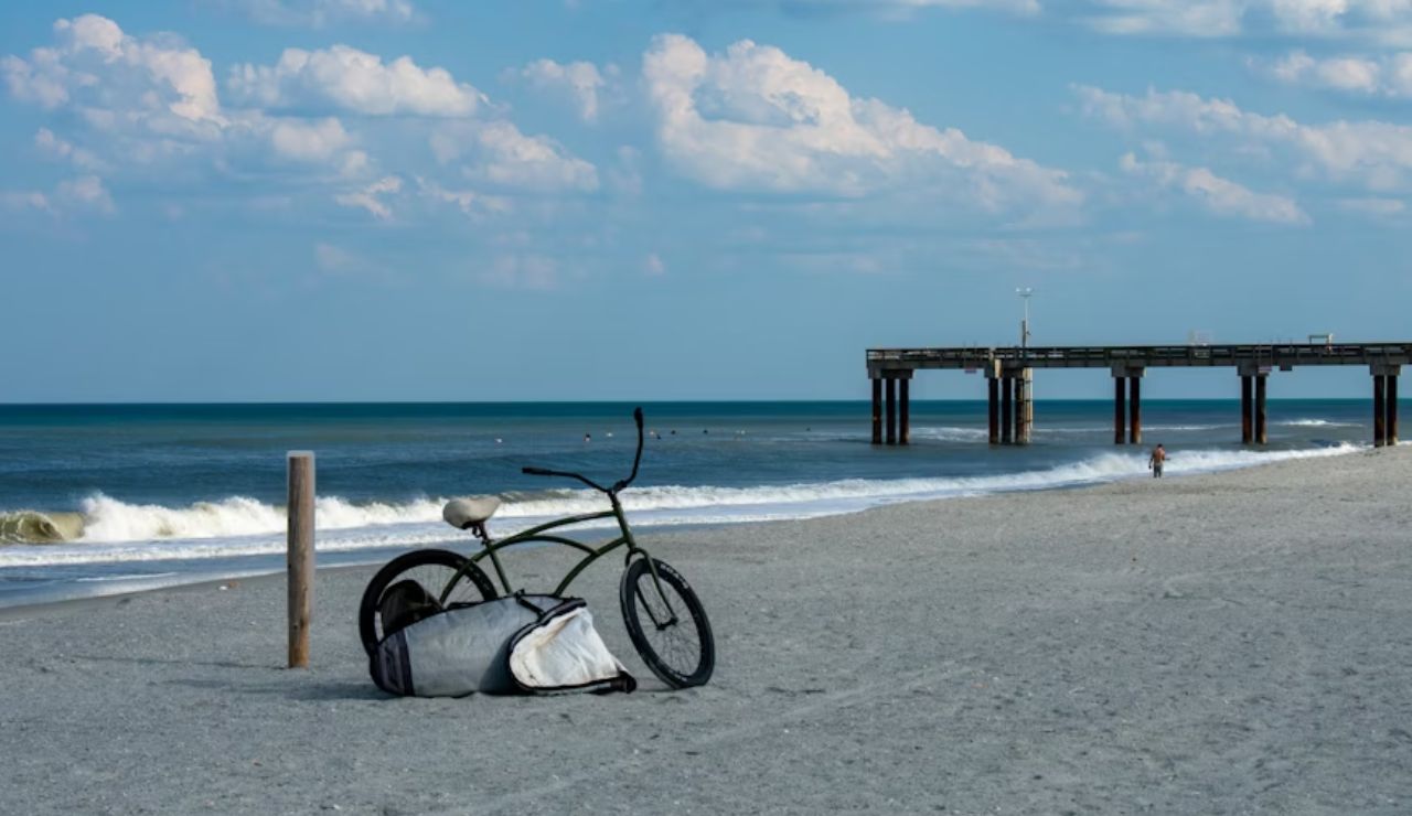 A lone bicycle with a bag rests on a quiet sandy beach near a pier under a blue sky with scattered clouds.