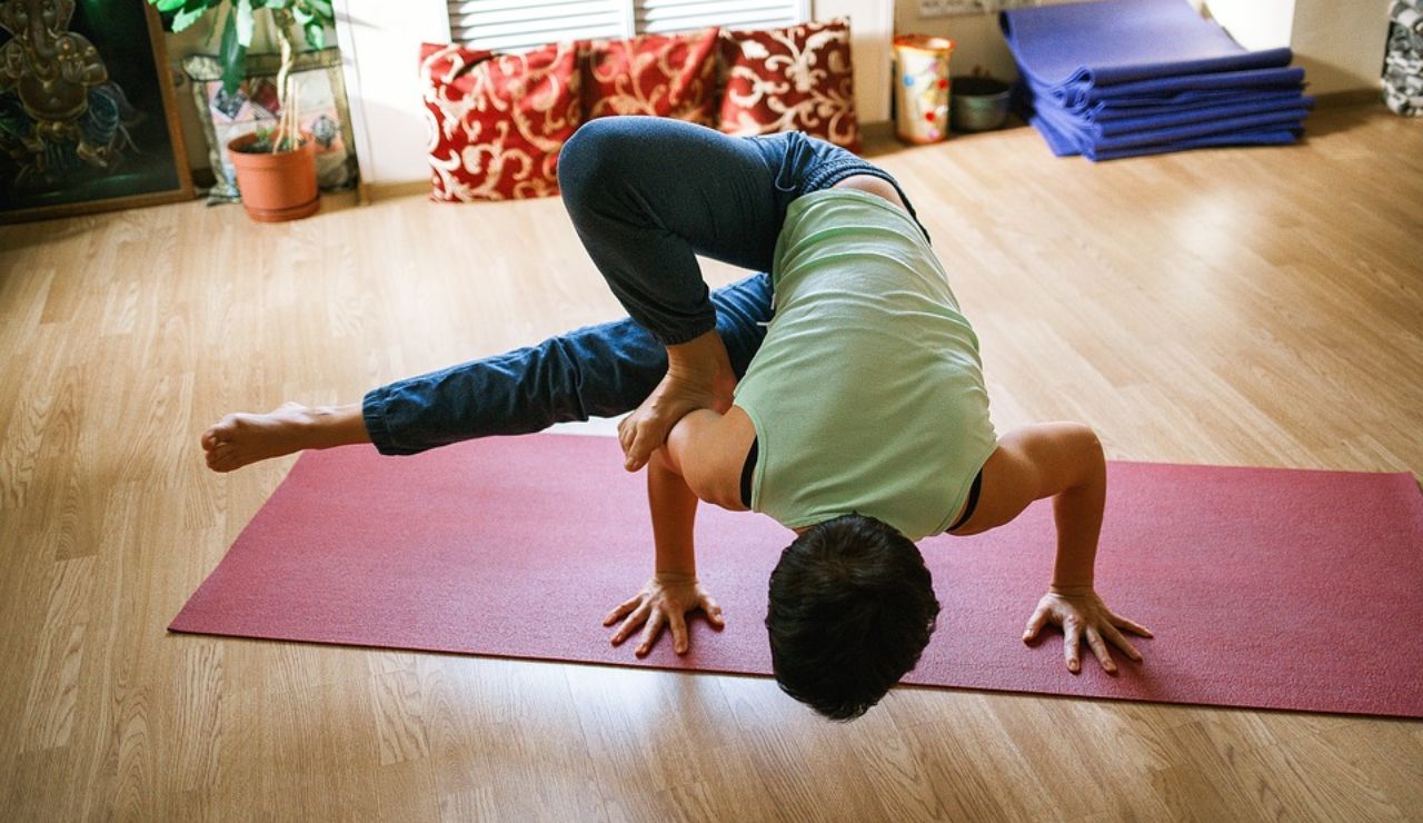 Person performing an advanced yoga pose on a red mat in a cozy indoor space with wooden flooring and plants.
