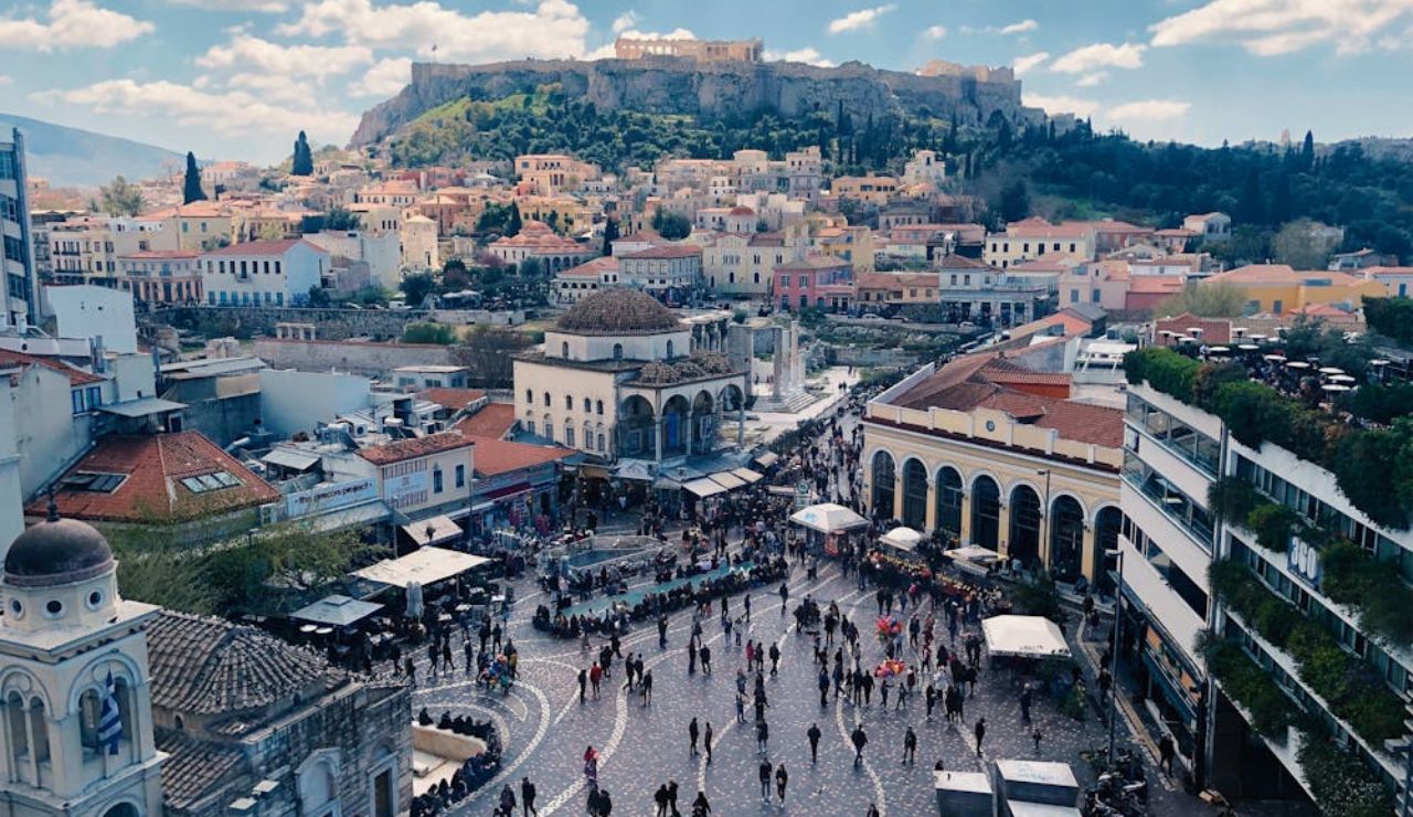 Bustling city square in Athens with crowds of people, historic buildings, and the Acropolis visible in the background on a sunny day.
