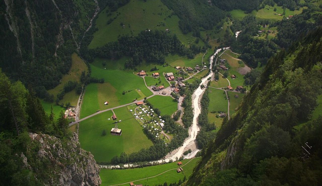 Aerial view of a peaceful mountain valley with green fields, scattered houses, and a river winding through the center, surrounded by forested cliffs.