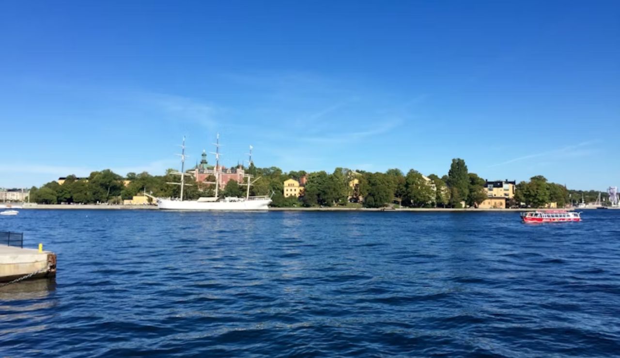 A white sailing ship and red tour boat glide across Stockholm’s blue harbor under clear skies.