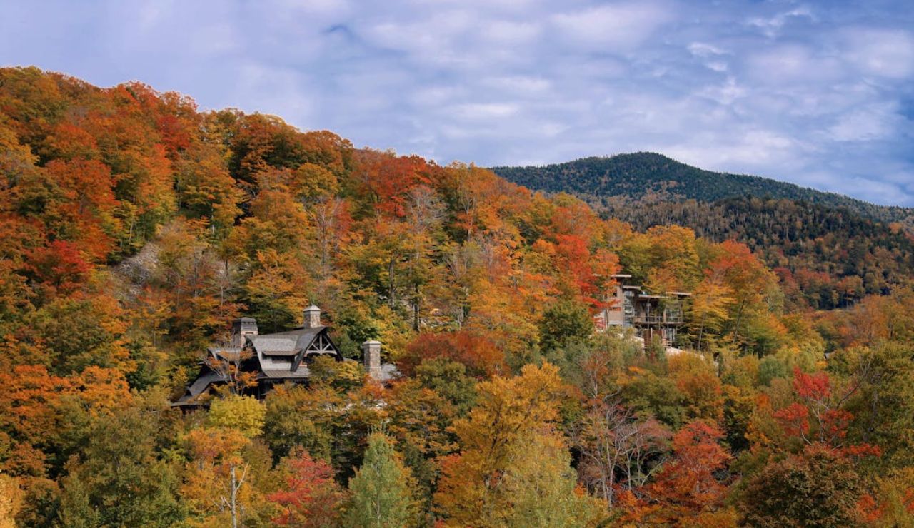 Houses nestled in a forested hillside with vibrant autumn foliage and mountain backdrop under a partly cloudy sky.
