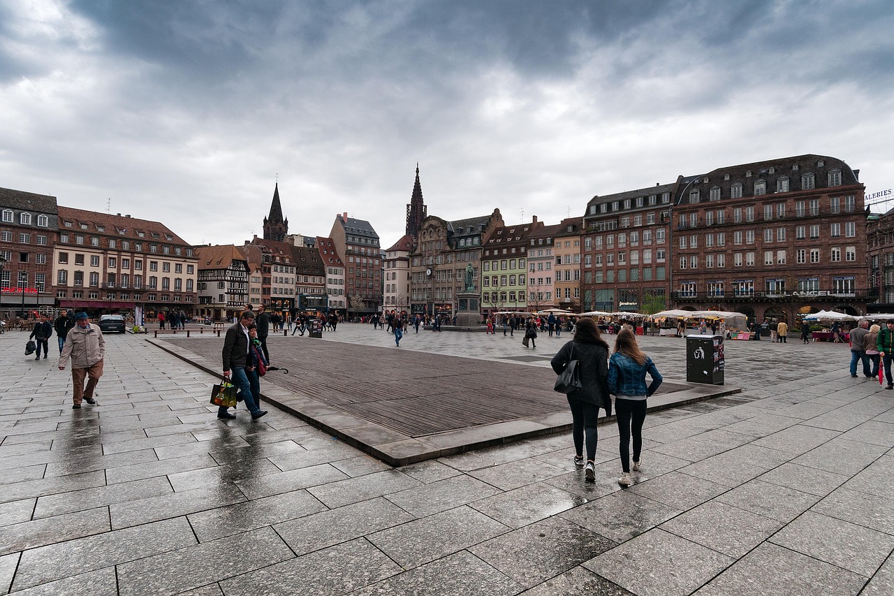 People walking across the wet cobblestone square of Place Kléber in Strasbourg, France, surrounded by historic buildings under a cloudy sky.