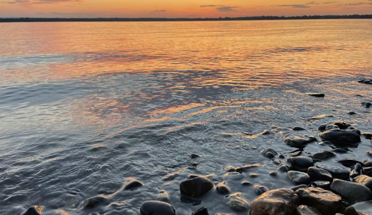 Rocky shoreline at sunset with calm water reflecting orange and pink hues across the surface.